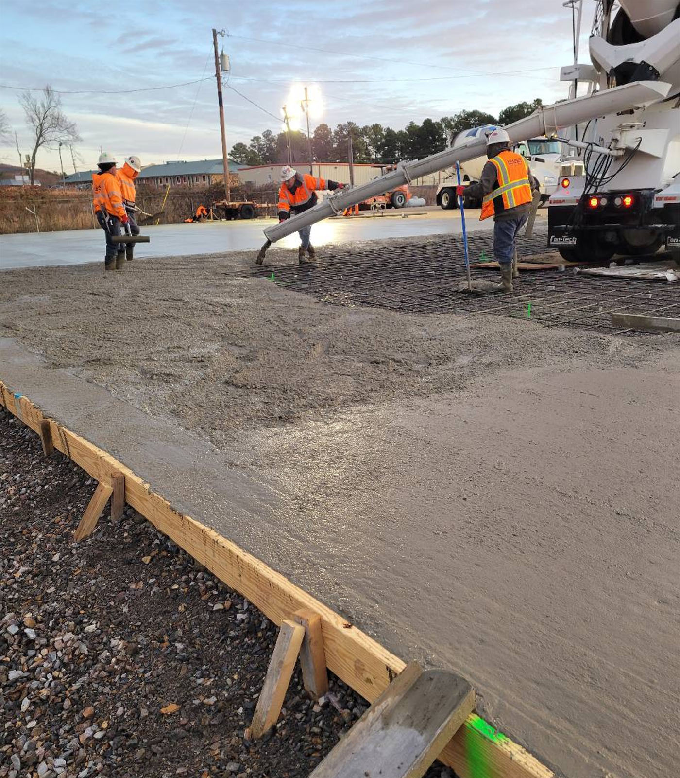 Construction crew in high-visibility vests pouring concrete from cement truck chute onto reinforced steel mesh with wooden forms defining the pour area
