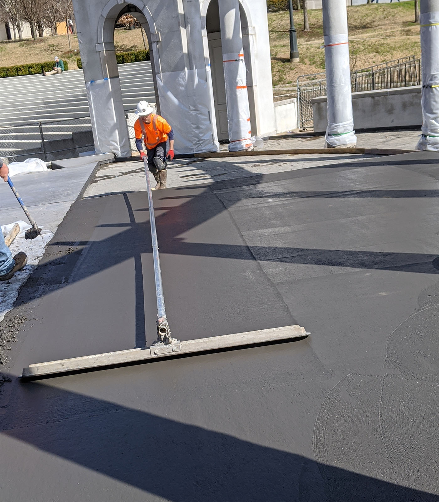 Construction worker in orange safety vest using long-handled screed board to level fresh concrete surface with precast stadium seating sections visible in background