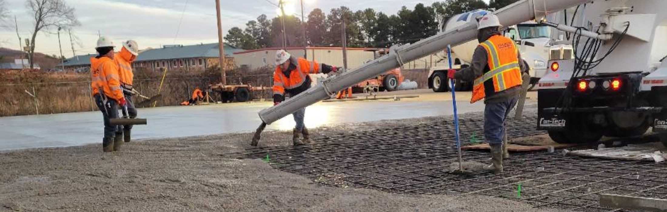 Backus Concrete construction workers in high-visibility vests pouring concrete from a cement truck chute onto a work site, with industrial buildings visible in the background